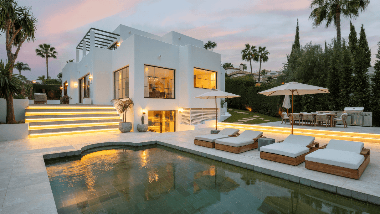 Modern white villa for sale in Nueva Andalucia with illuminated outdoor areas, private pool, sun loungers, and lush garden surroundings